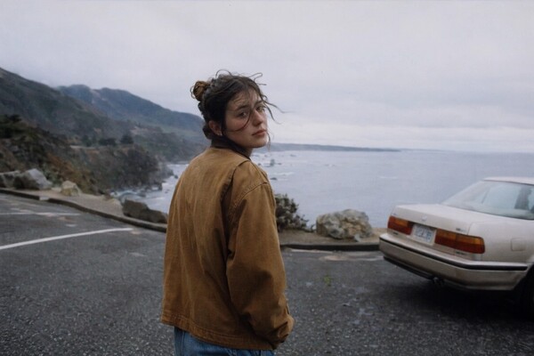 GPT Image 2 example: woman in canvas jacket looking back on a coastal overlook beside a vintage sedan