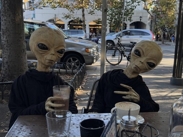 GPT Image 2 example: two alien figures in hoodies at an outdoor cafe with milkshake and city street background