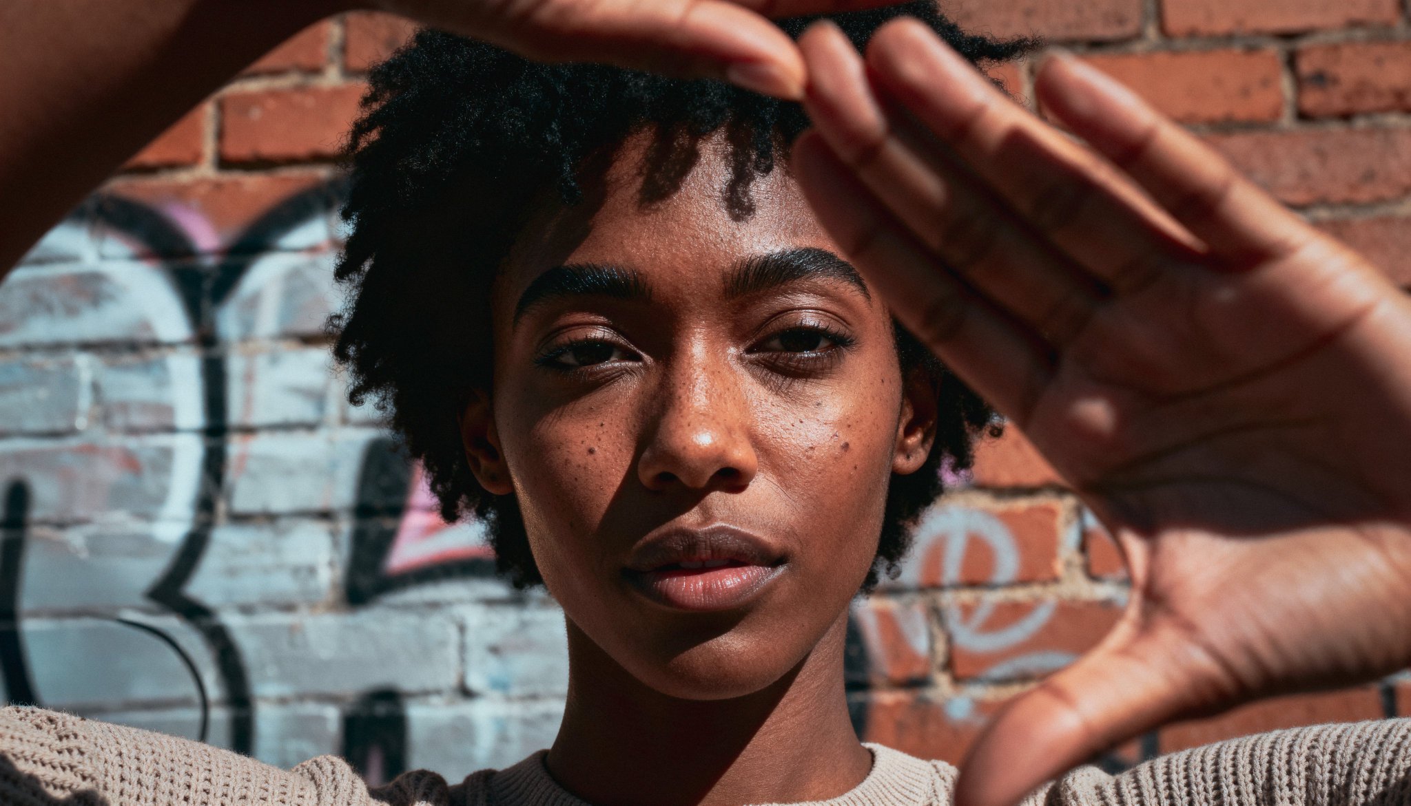 Seedream 4.5 example: portrait of a woman framing her face with hands like a camera viewfinder against brick graffiti